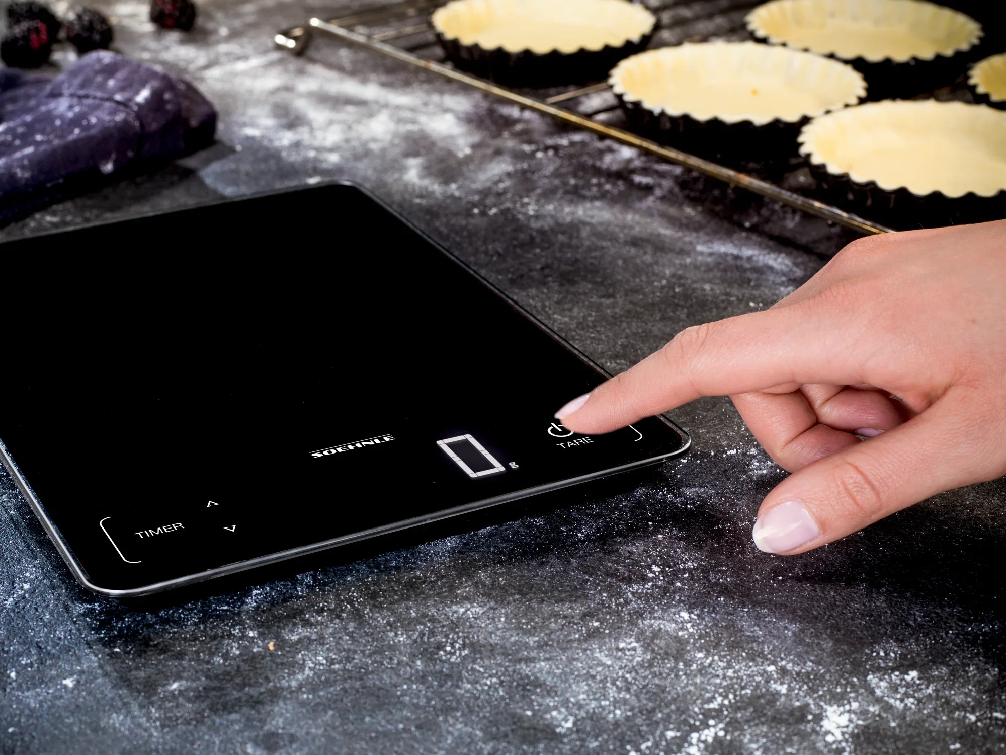 Een hand bedient de Page Profi 100 digitale keukenweegschaal op een werkoppervlak. Op de achtergrond zijn deegvormen en bloem te zien, wat duidt op een bakbereiding.