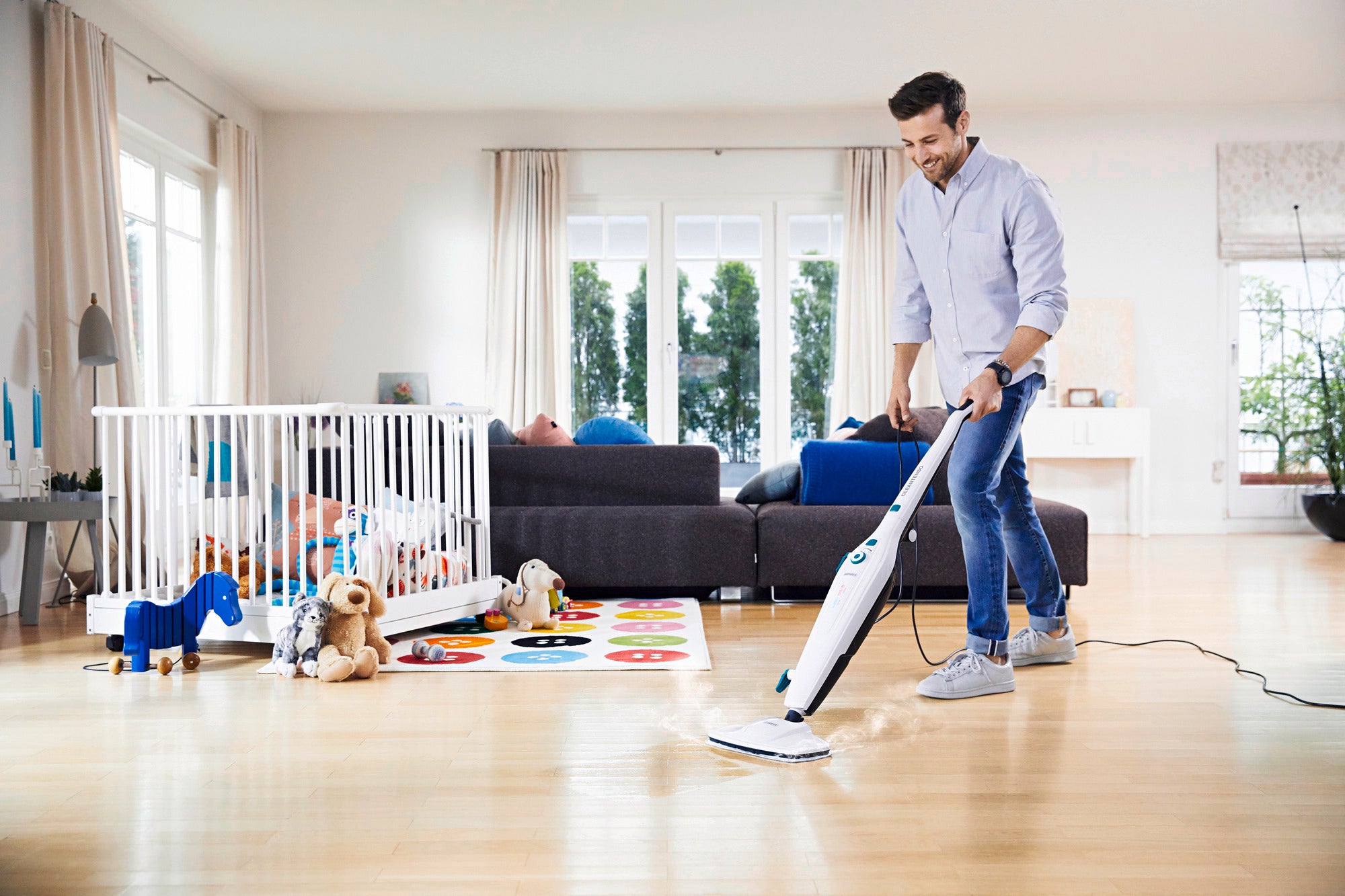 Un homme nettoie un parquet clair dans un salon moderne à l'aide du nettoyeur vapeur Clean Tenso. On aperçoit un parc pour bébé et des jouets à l'arrière-plan.