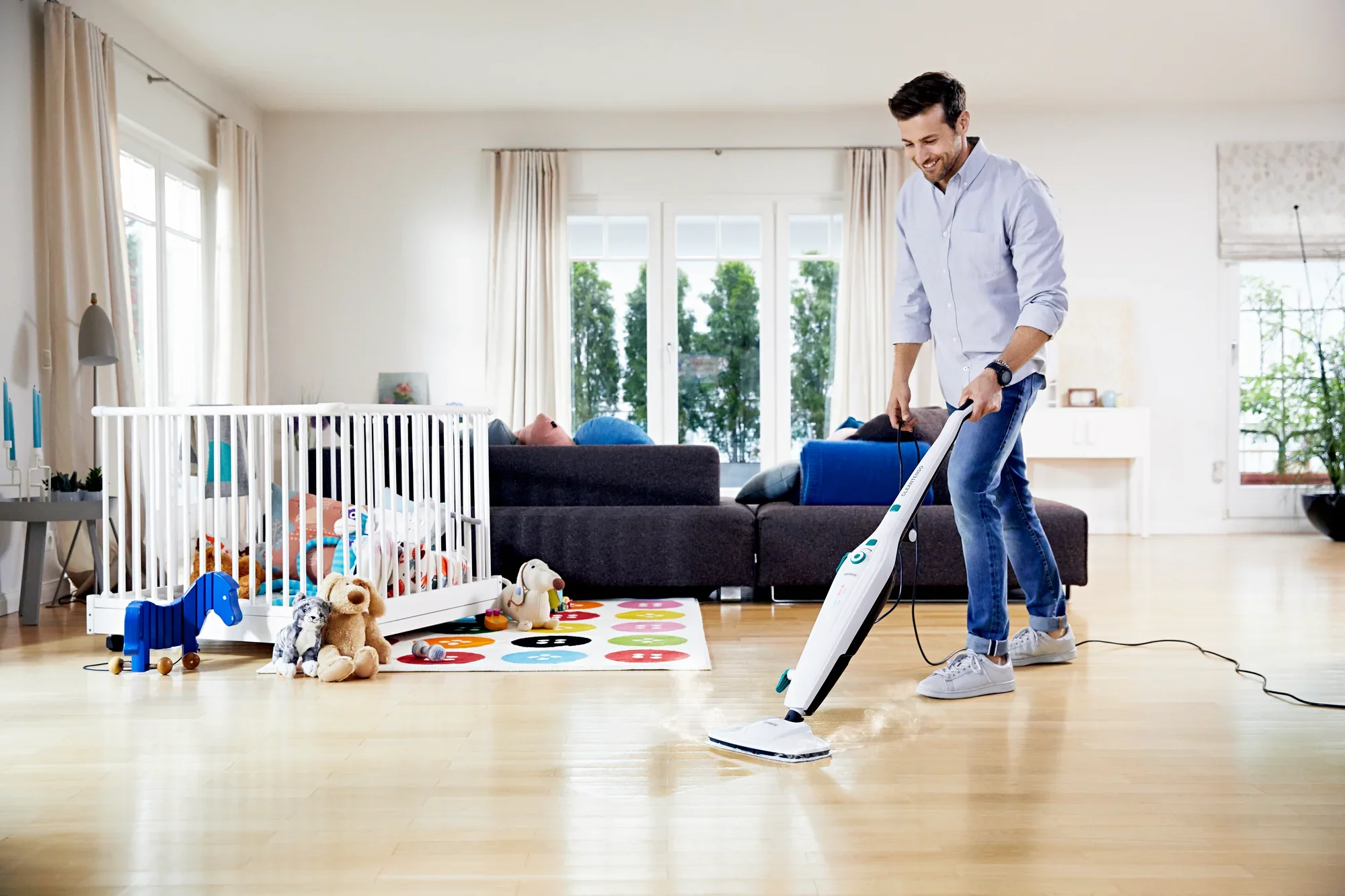 Un hombre limpia un suelo de madera clara en una moderna sala de estar con el limpiador a vapor Clean Tenso. Al fondo se ve un parque infantil y juguetes.