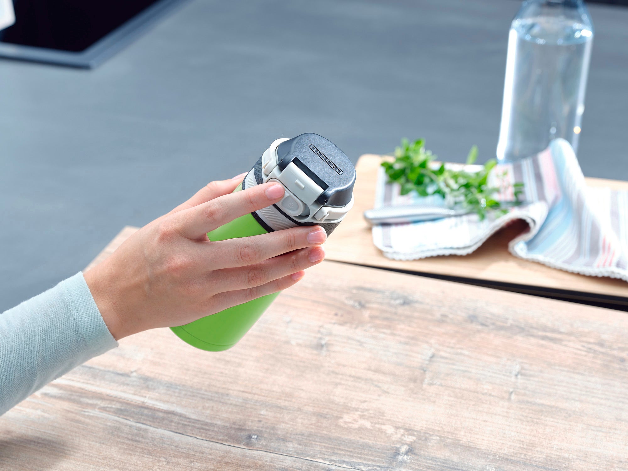 Eine Hand hält einen grünen Isolierbecher Flip mit 350 ml Fassungsvermögen. Im Hintergrund sind ein Geschirrtuch, frische Kräuter und eine Wasserflasche auf einem Holztisch zu sehen.