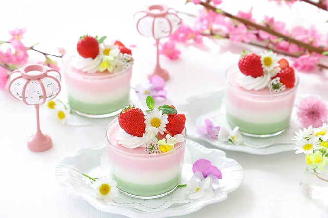 Leckeres Himbeer-Sahne-Dessert für die Frühlingszeit Dreifarbige Desserts in Glaesern mit Erdbeeren, Sahne und Blumen auf weissen Tellern, umgeben von rosa Blueten und dekorativen Lampen.