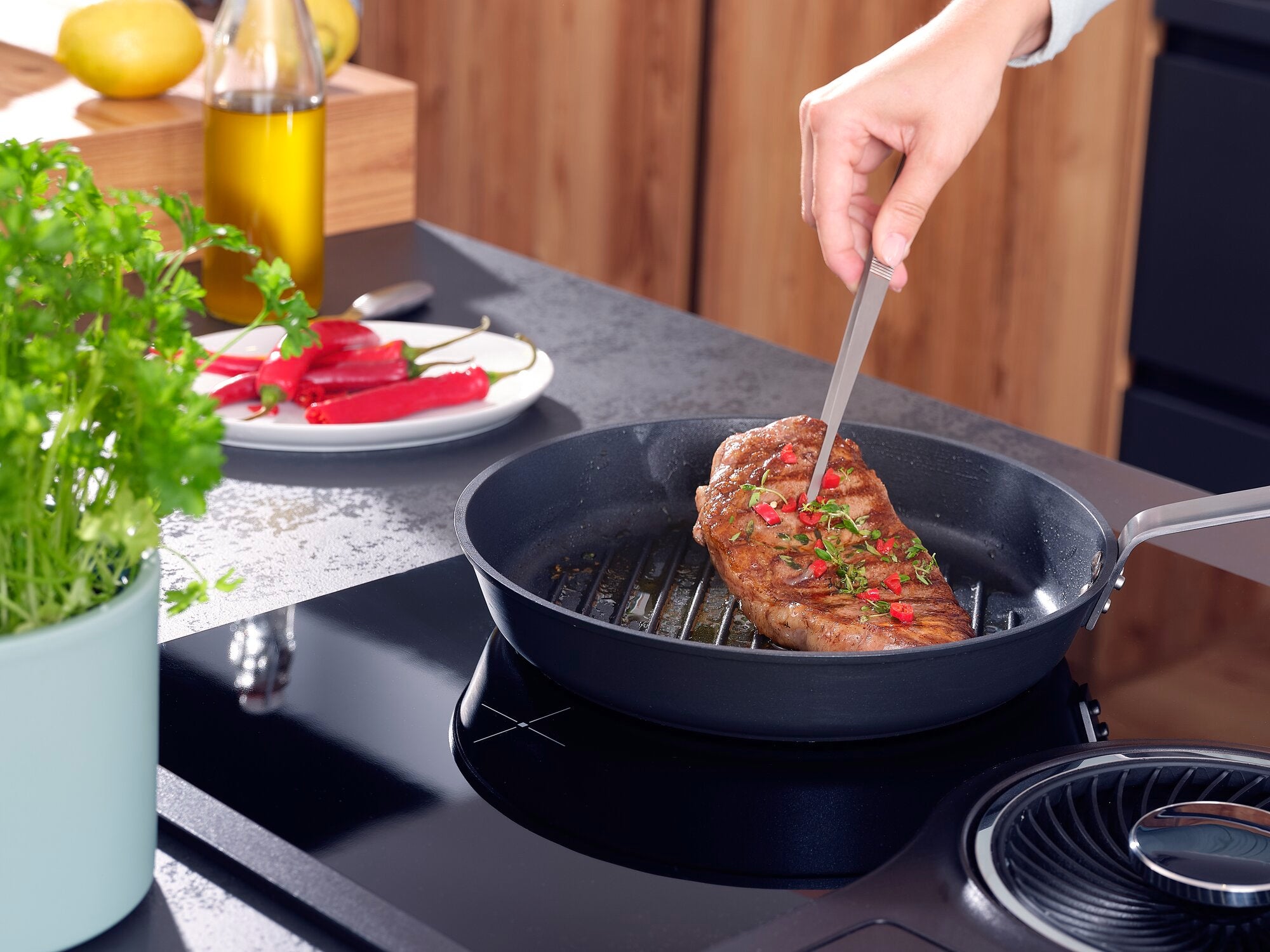Eine Hand hält eine Küchen-Pinzette und wendet ein saftiges Steak in einer Pfanne. Im Hintergrund sind frische Kräuter, Paprika und Olivenöl zu sehen.