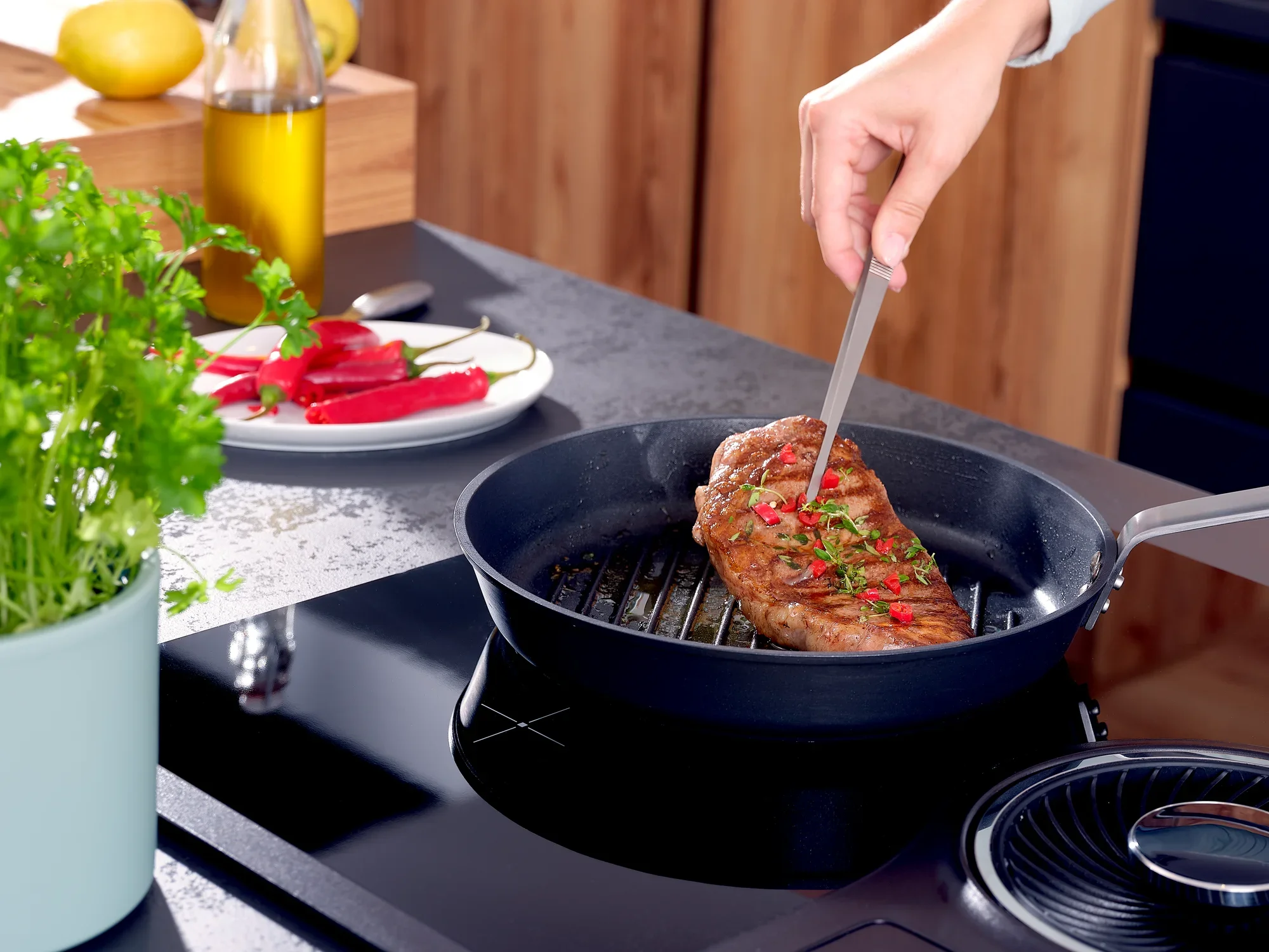 Een hand houdt een keukenpincet vast en draait een sappige biefstuk in een pan. Op de achtergrond zijn verse kruiden, paprika's en olijfolie te zien.