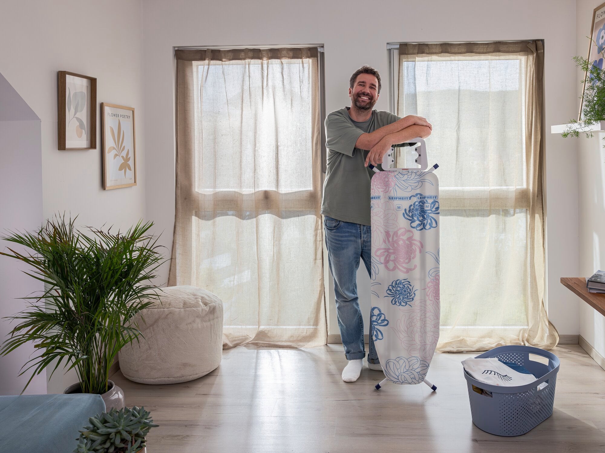 Ein Mann steht in einem hellen Raum und lehnt sich lächelnd an einen Bügeltisch Classic M Basic. Der Tisch hat ein florales Muster. Im Hintergrund sind Fenster mit Vorhängen und Pflanzen zu sehen.
