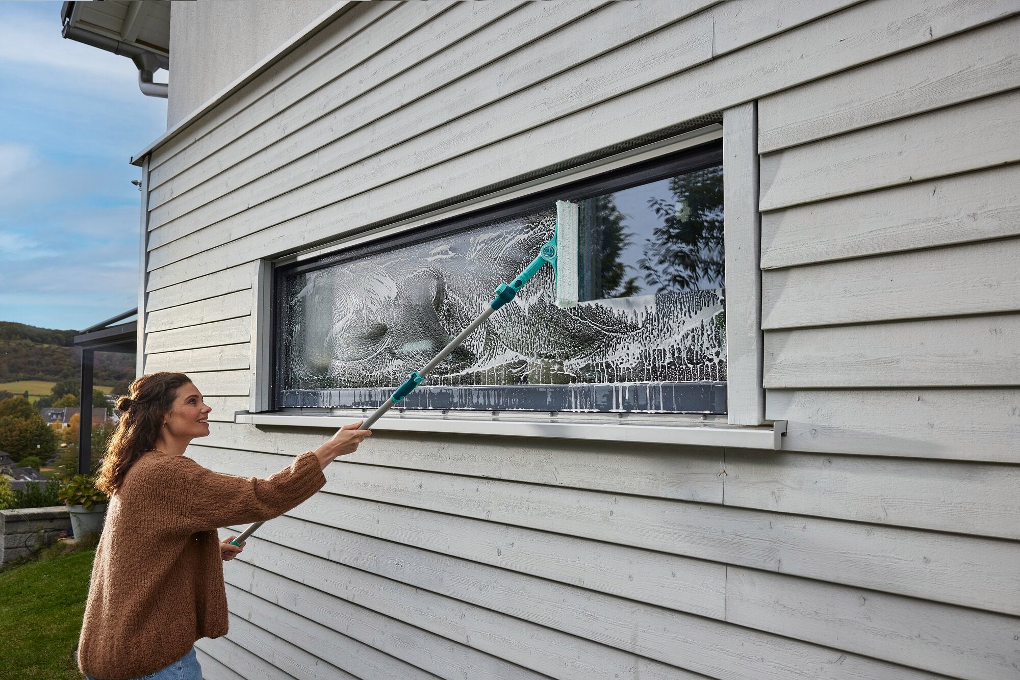Een vrouw maakt een groot raam schoon met de 4in1 telescopische raam- en kozijnreiniger. De reiniger bereikt moeiteloos hoge plekken en zorgt voor streeploos schoon.