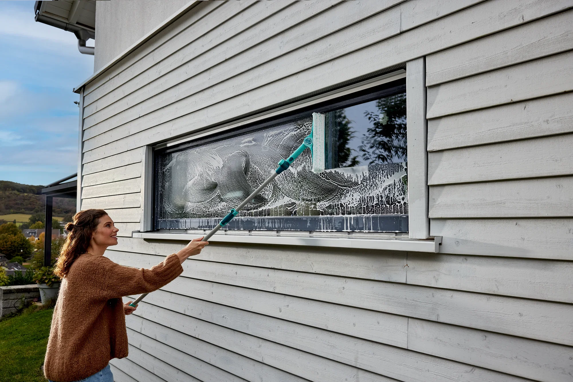 Een vrouw maakt een groot raam schoon met de 4in1 telescopische raam- en kozijnreiniger. De reiniger bereikt moeiteloos hoge plekken en zorgt voor streeploos schoon.