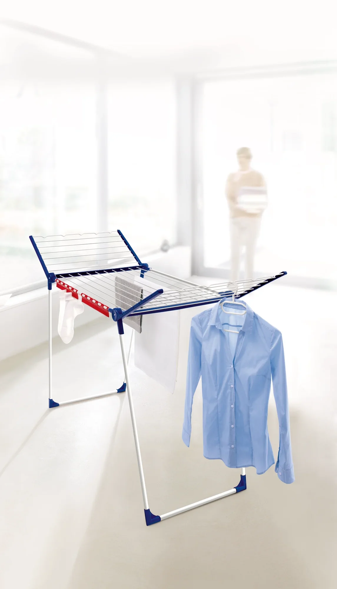 Suszarka Pegasus 200 Solid w jasnym pomieszczeniu. Na suszarce rozwieszone są niebieskie ubrania i ręczniki. W tle stoi jakaś osoba.