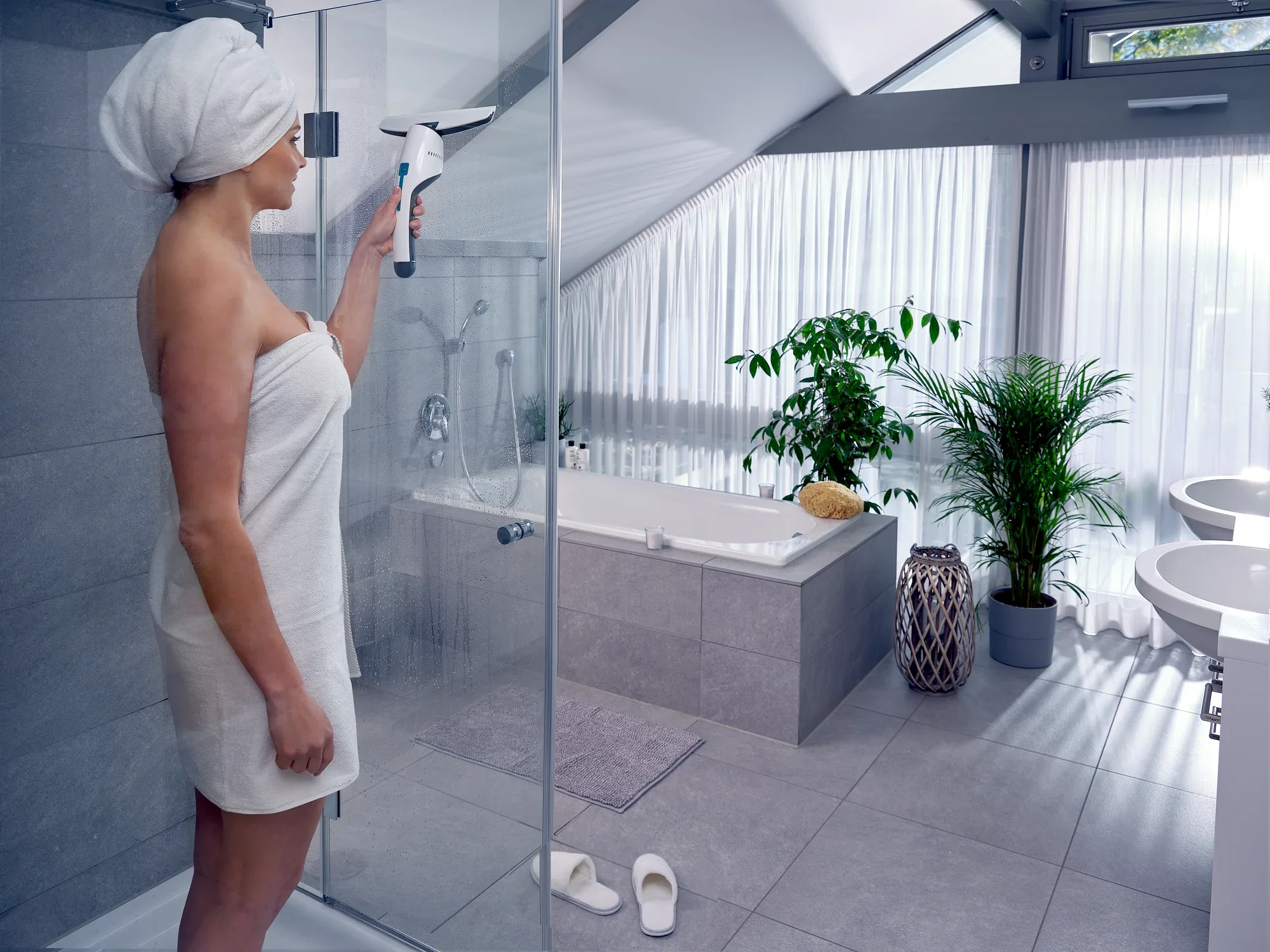 Eine Frau in einem modernen Badezimmer verwendet den Nemo Fenster- und Badsauger, um die Duschkabine zu reinigen. Der Raum ist hell und stilvoll eingerichtet mit Pflanzen und einer Badewanne.