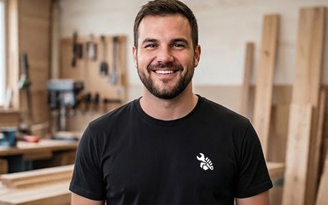 Ein lächelnder Tischler in seiner Werkstatt, der ein schwarzes ASTA Workwear T-Shirt mit einem gestickten Logo trägt